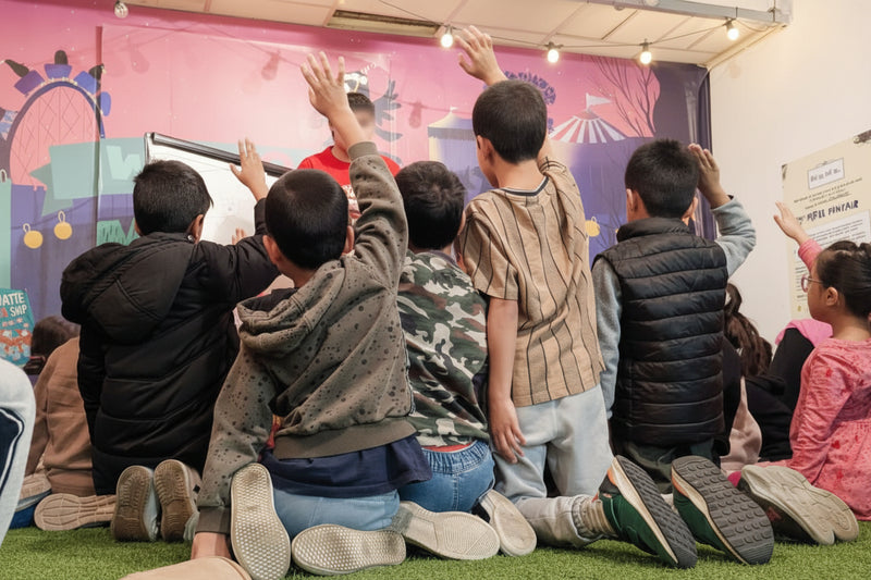 Children sitting on the floor with raised hands in front of a colorful wall.